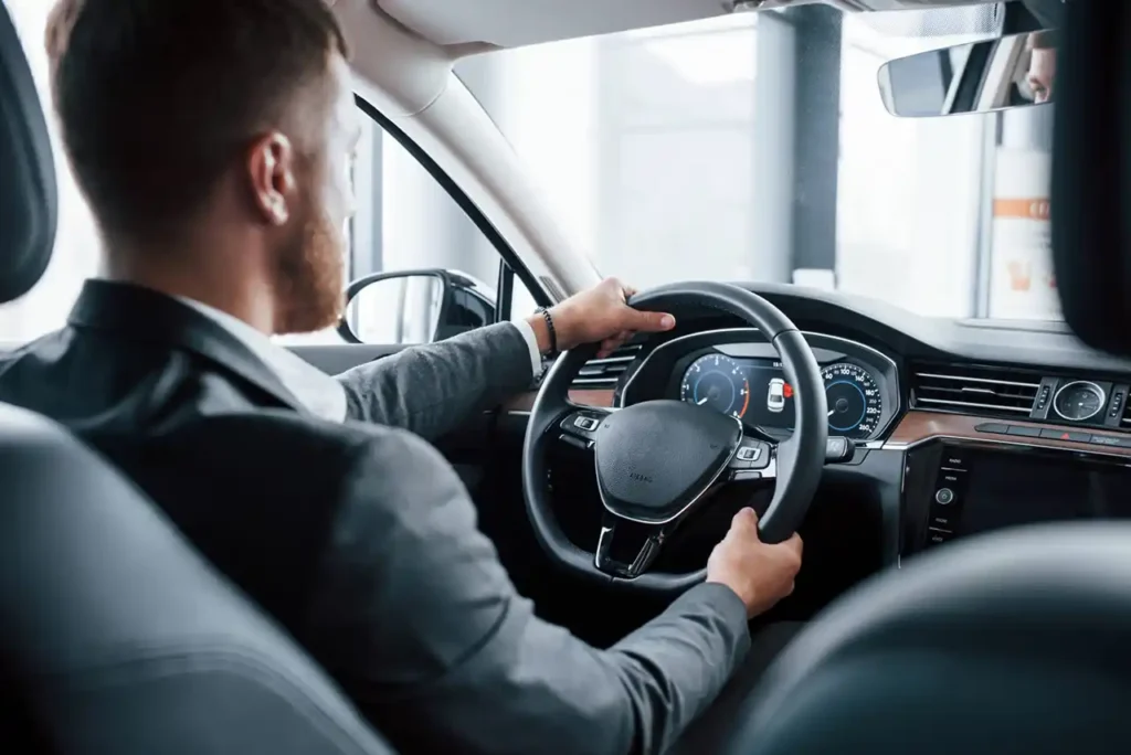 A uniformed chauffeur, wearing a professional suit and mask, is seated behind the wheel of a black Mercedes-Benz S-Class, preparing for a professional Pomona to and from LAX transfer.